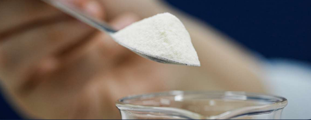A close-up of a spoonful of white powder being held above a glass container, with blurred hands and a deep blue background.