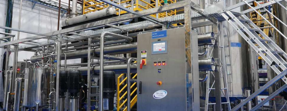 Industrial processing equipment featuring pipes, tanks, and a control panel, set in a spacious factory environment.