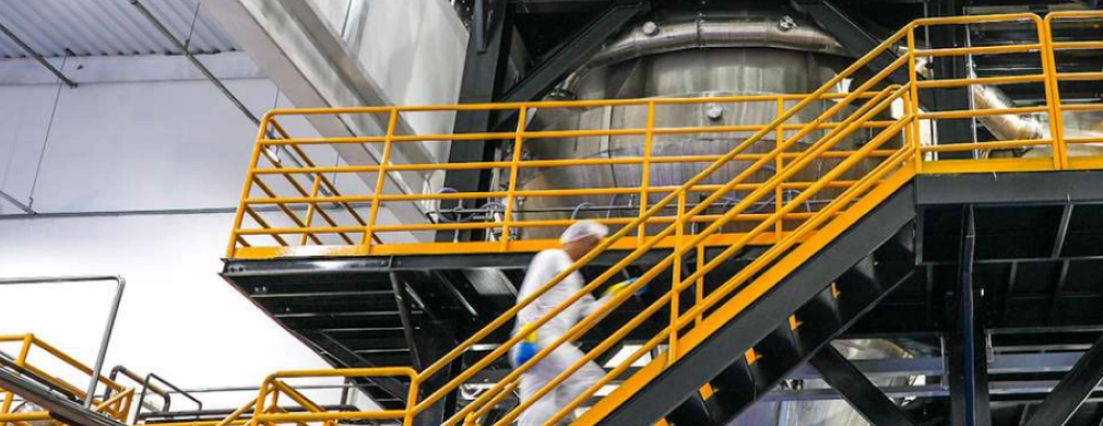 A worker in a white suit ascends a metal staircase with yellow railings in an industrial facility, leading to a large equipment chamber.