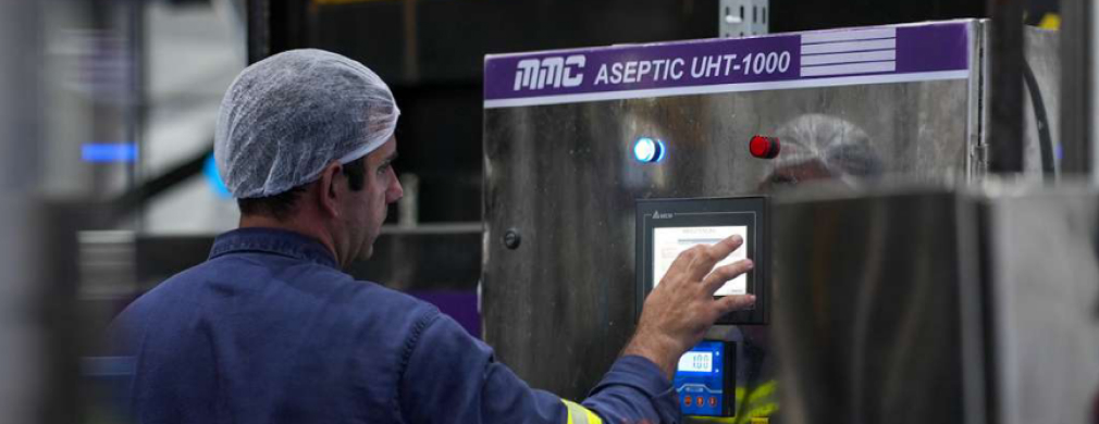 A worker operates a control panel of the Aseptic UHT-1000 machine in a sterile industrial environment, wearing a hairnet and blue uniform.
