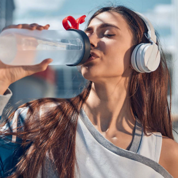 A woman in athletic attire drinks water from a clear bottle while wearing headphones, enjoying a sunny day outdoors.